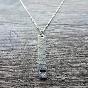May include: A silver-colored necklace with a textured, rectangular pendant. The pendant has a heart-shaped cutout near the bottom and hangs from a delicate chain. The background is a wooden surface.