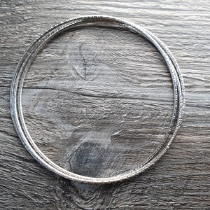 K&ouml;nnte beinhalten: Ein Set aus drei silbernen Armreifen mit strukturierter Oberfl&auml;che. Die Armreifen liegen &uuml;bereinander auf einer Holzoberfl&auml;che.