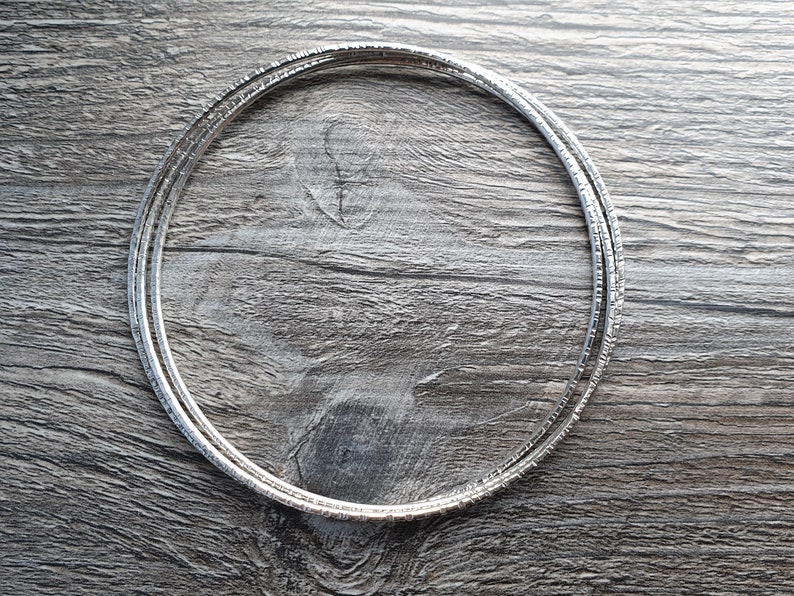 K&ouml;nnte beinhalten: Ein Set aus drei silbernen Armreifen mit strukturierter Oberfl&auml;che. Die Armreifen liegen &uuml;bereinander auf einer Holzoberfl&auml;che.