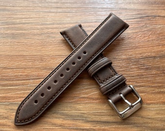 May include: A brown leather watch strap with a silver buckle. The strap has a stitched edge and a row of holes for adjusting the size.