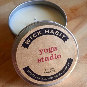 May include: A silver tin candle with a brown label that reads "WICK HABIT yoga studio Est. 2015 Austin, TX HAND-POURED SOY WAX CANDLES".