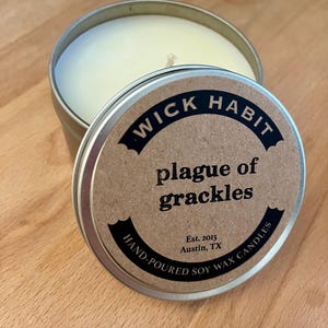 May include: A round tin candle with a brown label that reads "WICK HABIT plague of grackles Est. 2015 Austin, TX HAND-POURED SOY WAX CANDLES".
