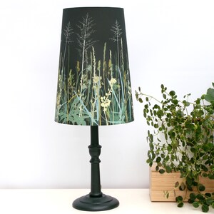 K&ouml;nnte beinhalten: Eine schwarze Tischlampe mit einem konischen Lampenschirm mit einem gr&uuml;nen und wei&szlig;en Blumendruck aus hohen Gr&auml;sern und Wildblumen. Der Lampenfu&szlig; ist dunkelgr&uuml;n.