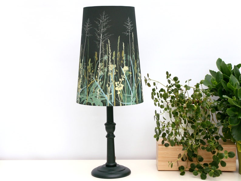 K&ouml;nnte beinhalten: Eine schwarze Tischlampe mit einem konischen Lampenschirm mit einem gr&uuml;nen und wei&szlig;en Blumendruck aus hohen Gr&auml;sern und Wildblumen. Der Lampenfu&szlig; ist dunkelgr&uuml;n.