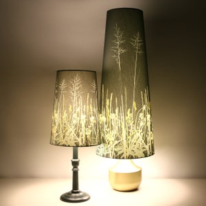 K&ouml;nnte beinhalten: Zwei Lampen mit hohen, zylindrischen Schirmen, die einen botanischen Druck aus gr&uuml;nen und gelben Gr&auml;sern und Blumen auf einem dunkelgrauen Hintergrund zeigen. Die kleinere Lampe hat einen schwarzen Fu&szlig;, w&auml;hrend die gr&ouml;&szlig;ere Lampe einen goldenen Fu&szlig; hat.