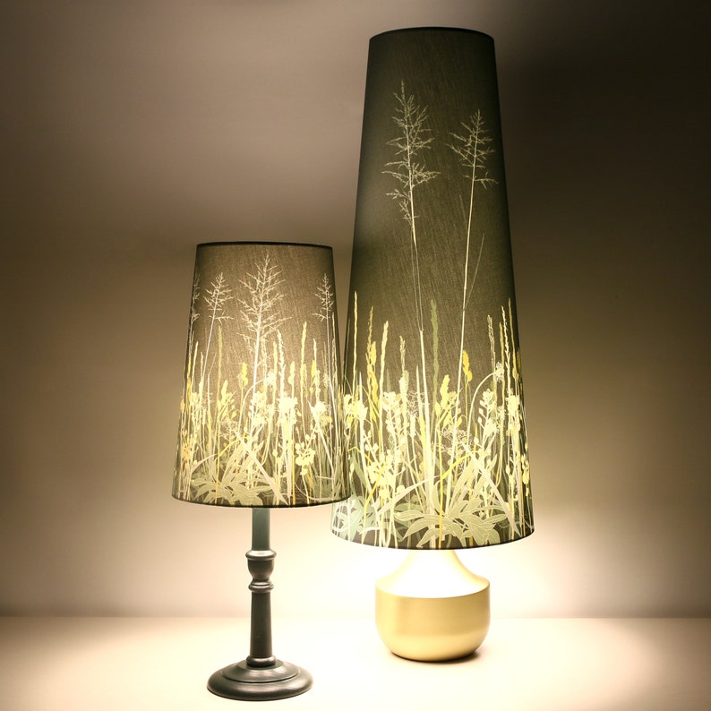 K&ouml;nnte beinhalten: Zwei Lampen mit hohen, zylindrischen Schirmen, die einen botanischen Druck aus gr&uuml;nen und gelben Gr&auml;sern und Blumen auf einem dunkelgrauen Hintergrund zeigen. Die kleinere Lampe hat einen schwarzen Fu&szlig;, w&auml;hrend die gr&ouml;&szlig;ere Lampe einen goldenen Fu&szlig; hat.
