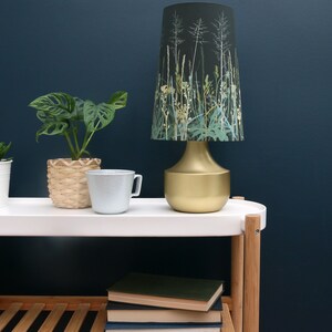 K&ouml;nnte beinhalten: Eine goldene Tischlampe mit einem schwarzen und gr&uuml;nen Lampenschirm mit floralem Muster. Die Lampe steht auf einem wei&szlig;en Regal mit einem Stapel B&uuml;cher und einer Topfpflanze.