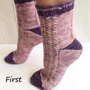May include: A pair of knitted socks in a pink and purple color scheme. The socks have a cable knit pattern and a contrasting purple heel and toe.