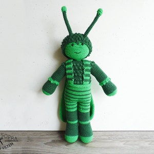May include: Green crocheted plush toy in the shape of a person wearing a green and black striped costume with long antennae. The toy is standing on a wooden surface.