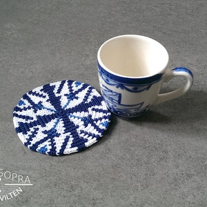 Könnte beinhalten: Ein blauer und weißer gehäkelter Untersetzer mit geometrischem Muster. Der Untersetzer liegt auf einer grauen Oberfläche neben einer weißen Keramiktasse mit blauen und weißen Blumendesigns.