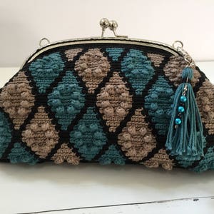 May include: A crocheted clutch bag with a black, brown, and teal diamond pattern. The bag has a silver clasp and a teal tassel with beads.