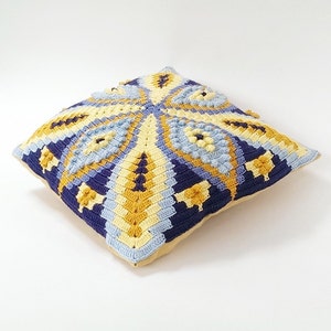 May include: A square crocheted cushion with a geometric pattern in shades of blue, yellow, and white. The cushion is made with a textured yarn and has a beige fabric backing.