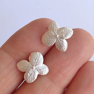 May include: A pair of silver earrings shaped like hydrangea flower blossoms. The earrings are delicate and have a textured surface.