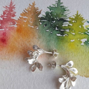 May include: Silver stud earrings with a botanical design. Each earring has a cluster of leaf-like shapes. The background is a watercolour painting of a forest with trees in shades of green, yellow, orange, and red.