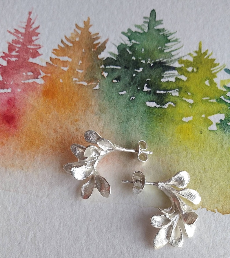 May include: Silver stud earrings with a botanical design. Each earring has a cluster of leaf-like shapes. The background is a watercolour painting of a forest with trees in shades of green, yellow, orange, and red.