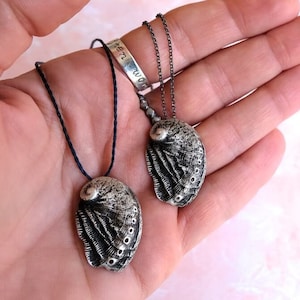Könnte beinhalten: Zwei silberne Halsketten mit Anhängern aus Abalone-Muscheln. Die Muscheln sind detailliert und haben eine strukturierte, gealterte Optik. Eine Halskette hat eine schwarze Schnur und die andere eine silberne Kette.