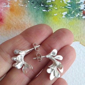 May include: A pair of silver earrings with a floral design. Each earring has a curved shape, with multiple leaf-like elements. The earrings have a post and butterfly clasp. The background is a watercolour painting with green, yellow, and orange hues.
