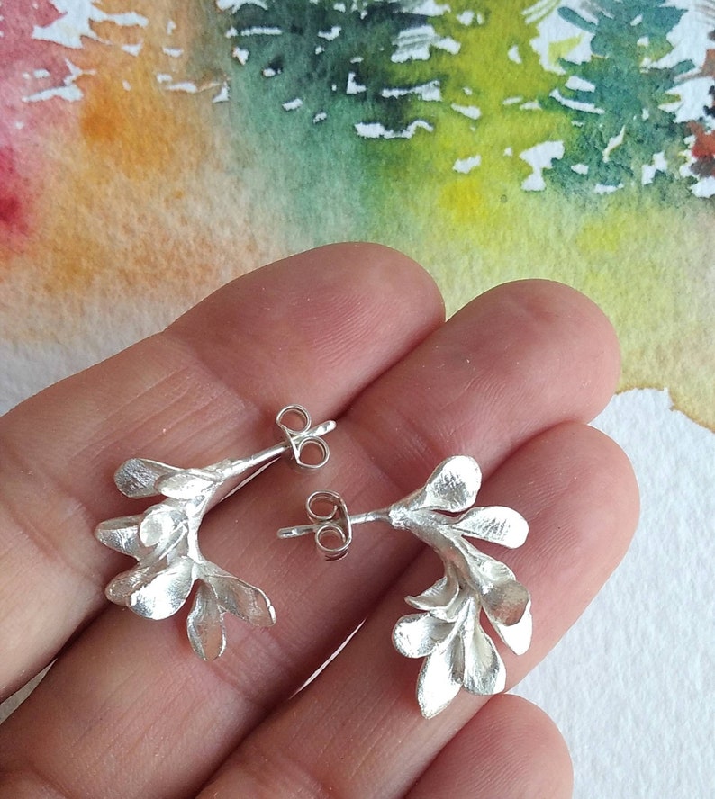 May include: A pair of silver earrings with a floral design. Each earring has a curved shape, with multiple leaf-like elements. The earrings have a post and butterfly clasp. The background is a watercolour painting with green, yellow, and orange hues.