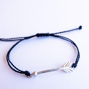 Può includere: Un braccialetto in corda nera con un ciondolo a forma di freccia d'argento. Il braccialetto è regolabile e può essere indossato al polso o alla caviglia.