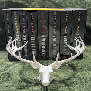 May include: A white deer head-shaped jewellery holder with two rings on the antlers. Behind it are several books with the titles "Throne of Glass", "Crown of Midnight", "Heir of Fire", "Queen of Shadows", "Empire of Storms", "Tower of Dawn", and "Kingdom of Ash" by Sarah J. Maas.