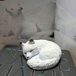 May include: A white arctic fox figurine curled up, with black-tipped ears, resting on a book. The book has visible text. The background shows a snowy landscape and another fox.