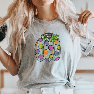 K&ouml;nnte beinhalten: Ein hellgraues T-Shirt mit einem bunten K&uuml;rbisdesign. Der K&uuml;rbis hat eine hellgr&uuml;ne Basis mit gro&szlig;en Punkten in Orange, Lila und Rosa. Der Stiel ist schwarz-wei&szlig; gestreift, mit gr&uuml;nen Bl&auml;ttern.