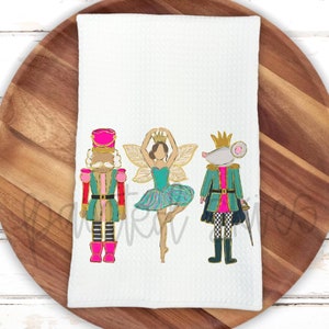 May include: A white kitchen towel with an embroidered design of a nutcracker, a ballerina, and a mouse king. The nutcracker is wearing a green and pink uniform, the ballerina is wearing a turquoise tutu, and the mouse king is wearing a green and pink uniform with a crown.