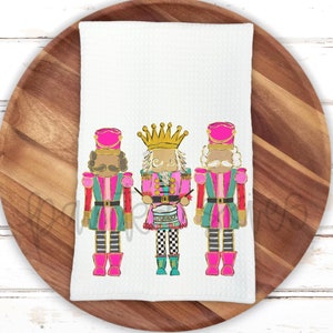 May include: Three pink and green nutcracker figurines with gold accents. The middle nutcracker is playing a drum. The nutcrackers are on a white kitchen towel.