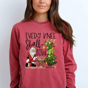 May include: Crimson long-sleeve shirt with the text "EVERY KNEE SHALL BOW" and a graphic of Santa Claus kneeling before a manger and a decorated Christmas tree. The shirt is a solid crimson color.