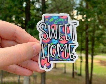 May include: A sticker featuring the outline of the state of Alabama with a floral pattern and the text "Sweet Home" in white lettering.