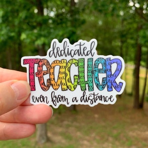 May include: A rainbow-colored sticker with the words "dedicated teacher even from a distance". The word "teacher" is decorated with a colorful pattern.