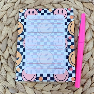 May include: A black and white checkered notepad with a white lined area for writing. The notepad is decorated with a repeating pattern of yellow smiley faces with pink outlines and small orange flowers.