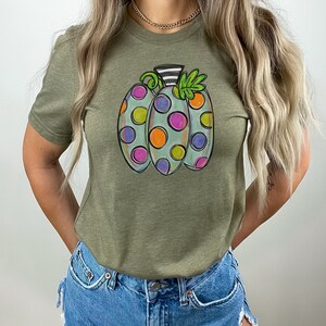 K&ouml;nnte beinhalten: Olivgr&uuml;nes T-Shirt mit einem skurrilen K&uuml;rbisdesign. Der K&uuml;rbis ist hellblau mit bunten Tupfen in Lila, Orange und Gr&uuml;n. Er hat einen schwarz-wei&szlig; gestreiften Stiel und gr&uuml;ne Bl&auml;tter.
