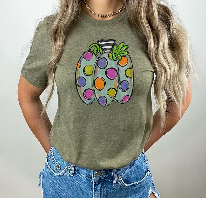 K&ouml;nnte beinhalten: Olivgr&uuml;nes T-Shirt mit einem skurrilen K&uuml;rbisdesign. Der K&uuml;rbis ist hellblau mit bunten Tupfen in Lila, Orange und Gr&uuml;n. Er hat einen schwarz-wei&szlig; gestreiften Stiel und gr&uuml;ne Bl&auml;tter.