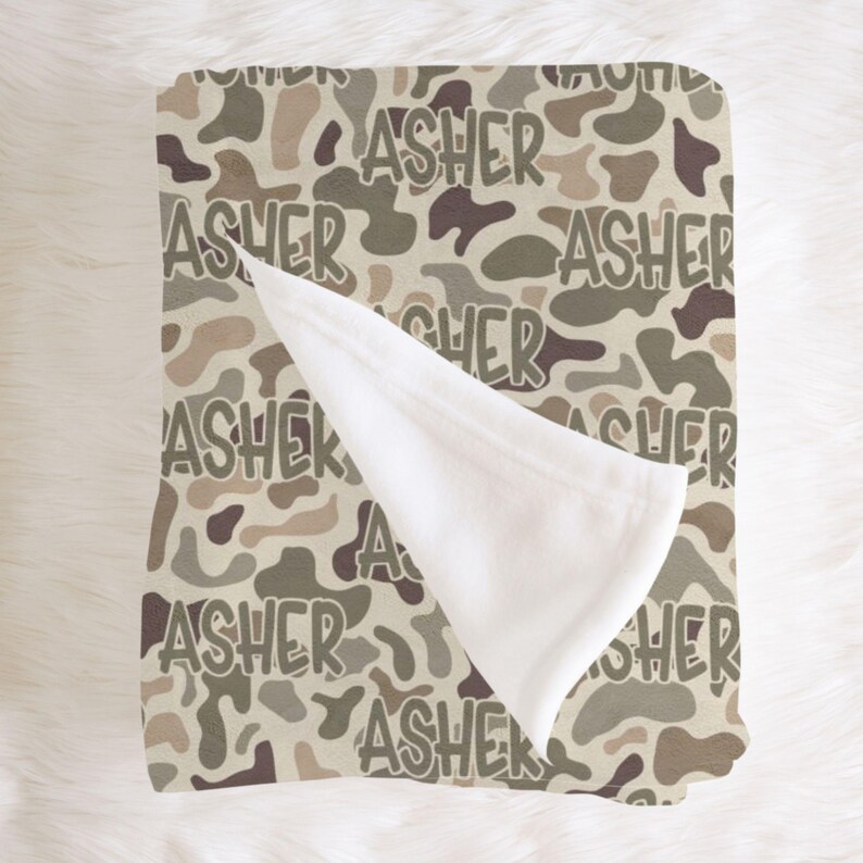 Op de afbeelding: Een babydeken met een camouflagepatroon in bruin, beige en groene tinten. De naam "ASHER" staat herhaaldelijk op de deken in een speels lettertype. Een deel van de deken is omgeslagen en toont een zachte, witte onderkant.