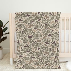 Op de afbeelding: Een beige babydeken met een camouflagepatroon in bruin, beige en grijstinten. De naam "ASHER" staat herhaaldelijk in het ontwerp. De deken is over een wieg gedrapeerd.