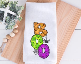 May include: White waffle-weave kitchen towel with the word "BOO" in orange, green, and purple bubble letters. A black spider and spiderweb accent the design, perfect for Halloween.