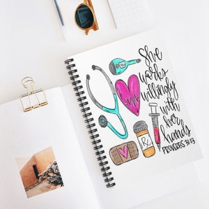 May include: A white spiral notebook with a colorful illustration of a stethoscope, a heart, a bandage, a pill bottle, and a syringe. The text "She works willingly with her hands Proverbs 31:13" is written in black cursive.