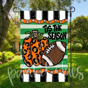 May include: A green, white, and black striped garden flag with an orange and black leopard print pumpkin and a brown football. The text "Tis the Season" is written in white on the flag.