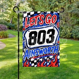 May include: A rectangular garden flag featuring a white license plate graphic with the number 803 in black. The text LET'S GO appears in red above the plate, and AINSWORTH appears in blue below. The background has red flames and a checkered pattern.