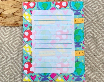 May include: A notepad with a lined paper overlay. The notepad has a teal background with a pattern of red apples, globes, pencils, and hearts. A ruler is printed on the side of the notepad.