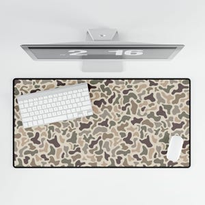 May include: A rectangular desk mat with a camouflage pattern in shades of brown, tan, and green. A white keyboard and mouse are on the mat. A computer monitor is above the mat.