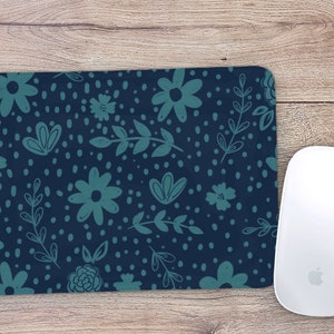 May include: A rectangular mousepad with a navy blue background and a teal floral pattern. The design includes flowers, leaves, and small dots. A white wireless mouse is positioned to the right of the mousepad.