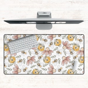 May include: A rectangular desk mat featuring a repeating pattern of bees, beehives, and flowers in shades of yellow, pink, and green on a white background. A white keyboard and mouse are on the mat, with a computer monitor above.
