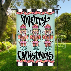 May include: A garden flag featuring three colorful nutcracker soldiers with the words "Merry Christmas" in a whimsical font. The flag has a light blue background with a black and white striped border.