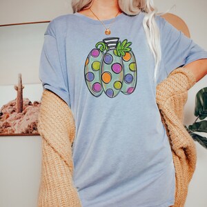 K&ouml;nnte beinhalten: Hellblaues T-Shirt mit einem bunten K&uuml;rbisdesign. Der K&uuml;rbis hat drei Abschnitte mit verschiedenfarbigen Punkten, die schwarz umrandet sind. Das Shirt wird mit einem beigefarbenen Cardigan getragen.