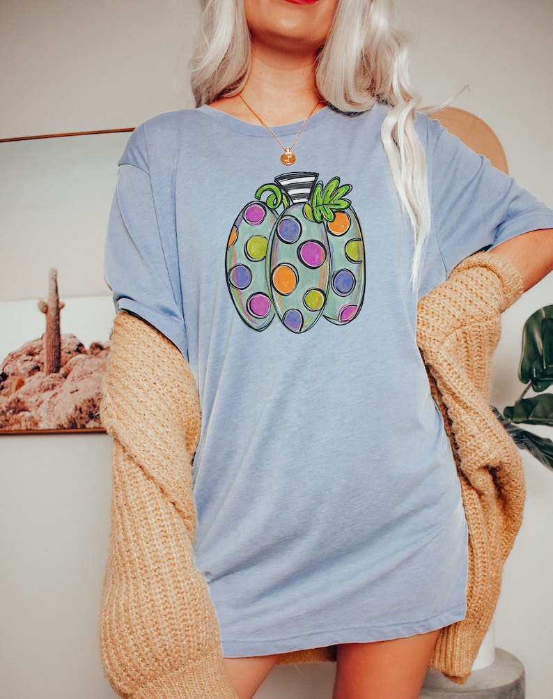 K&ouml;nnte beinhalten: Hellblaues T-Shirt mit einem bunten K&uuml;rbisdesign. Der K&uuml;rbis hat drei Abschnitte mit verschiedenfarbigen Punkten, die schwarz umrandet sind. Das Shirt wird mit einem beigefarbenen Cardigan getragen.