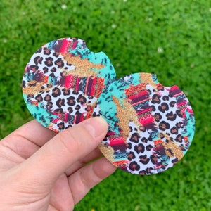 May include: Two round car coasters with a colorful design. The coasters feature a turquoise background with gold glitter accents, a leopard print, and a Southwestern-inspired pattern in red, pink, and brown.