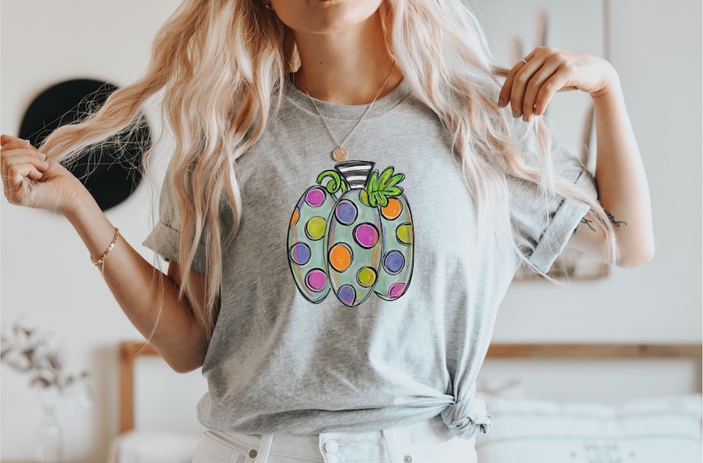 K&ouml;nnte beinhalten: Ein hellgraues T-Shirt mit einem bunten K&uuml;rbisdesign. Der K&uuml;rbis hat eine hellgr&uuml;ne Basis mit gro&szlig;en Punkten in Orange, Lila und Rosa. Der Stiel ist schwarz-wei&szlig; gestreift, mit gr&uuml;nen Bl&auml;ttern.