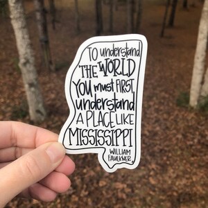 May include: A white sticker with a black outline of the state of Mississippi. The sticker features a quote by William Faulkner: "To understand the world you must first understand a place like Mississippi."